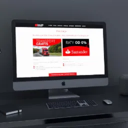 Ekran komputera iMac wyświetlający stronę internetową z promocjami transportu i rat 0%, na czarnym biurku z lampką, głośnikiem i akcesoriami.