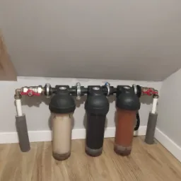 Trzy filtry wody o różnym stopniu zabrudzenia, zamontowane na rurach w pomieszczeniu ze skosami, ukazujące stopień filtracji wody.