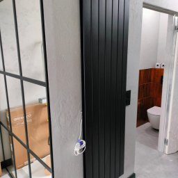 AQUA PLUS ANDRIY VORONOVSKIY - Czarny, pionowy grzejnik panelowy na szarej ścianie, widok z boku. W tle fragment łazienki z toaletą. Obok grzejnika widoczne przewody i skrzynka z szybami.