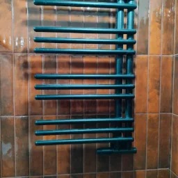 Czarny, drabinkowy grzejnik łazienkowy na ścianie z brązowych, błyszczących płytek w układzie pionowym. Widoczny fragment ściany i narożnik.