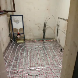 Białe rury ogrzewania podłogowego ułożone na srebrnej folii z czerwoną siatką w remontowanej łazience. Widoczny stelaż podtynkowy Grohe.