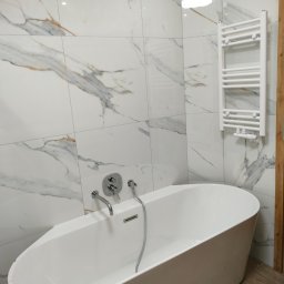 AQUA PLUS ANDRIY VORONOVSKIY - Biała, wolnostojąca wanna z chromowaną armaturą na tle marmurowych płytek i grzejnika drabinkowego w nowoczesnej łazience.