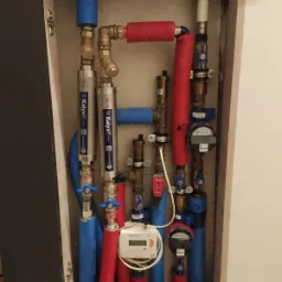 Węzeł cieplny w szafce: rury w izolacji czerwonej i niebieskiej, zawory, filtry marki Kalyxx i liczniki. Widoczne elementy instalacji wodnej i grzewczej.