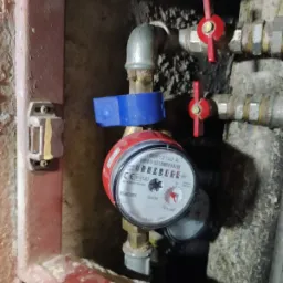 Zamontowany licznik wody z niebieską nakładką, widoczny na tle instalacji hydraulicznej z zaworami z czerwonymi uchwytami, umieszczony w zabudowie.