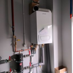 Andrzej Voronovski - Biały piec gazowy marki Broje zamontowany na szarej ścianie z widocznymi rurami miedzianymi i zaworami. Częściowo widoczne kartony i elementy instalacji.