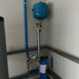 AQUA PLUS ANDRIY VORONOVSKIY - Niebieski zbiornik hydroforowy i filtr wody w pomieszczeniu technicznym, z widocznymi rurami i manometrem, na tle białych ścian i szarych rur instalacyjnych.