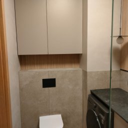 AQUA PLUS ANDRIY VORONOVSKIY - Nowoczesna łazienka w beżach: podwieszana toaleta z czarnym przyciskiem, szafki nad nią, pralka pod blatem i szklana kabina prysznicowa. Minimalistyczny design.