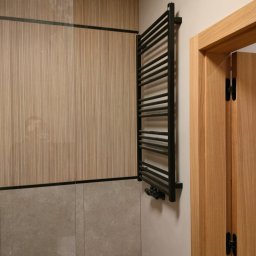 AQUA PLUS ANDRIY VORONOVSKIY - Czarny, drabinkowy grzejnik łazienkowy na ścianie z drewnianą okładziną i szarą płytką, obok drewniane drzwi z czarnymi zawiasami. Nowoczesne wykończenie łazienki.