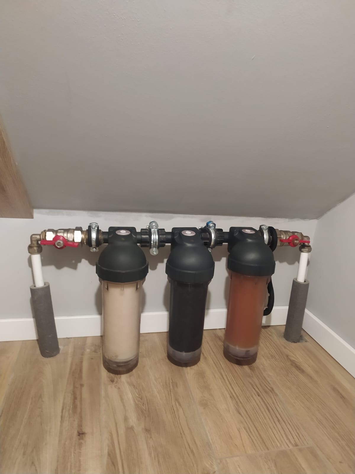 Trzy filtry wody o różnym stopniu zabrudzenia, zamontowane na rurach w pomieszczeniu ze skosami, ukazujące stopień filtracji wody.