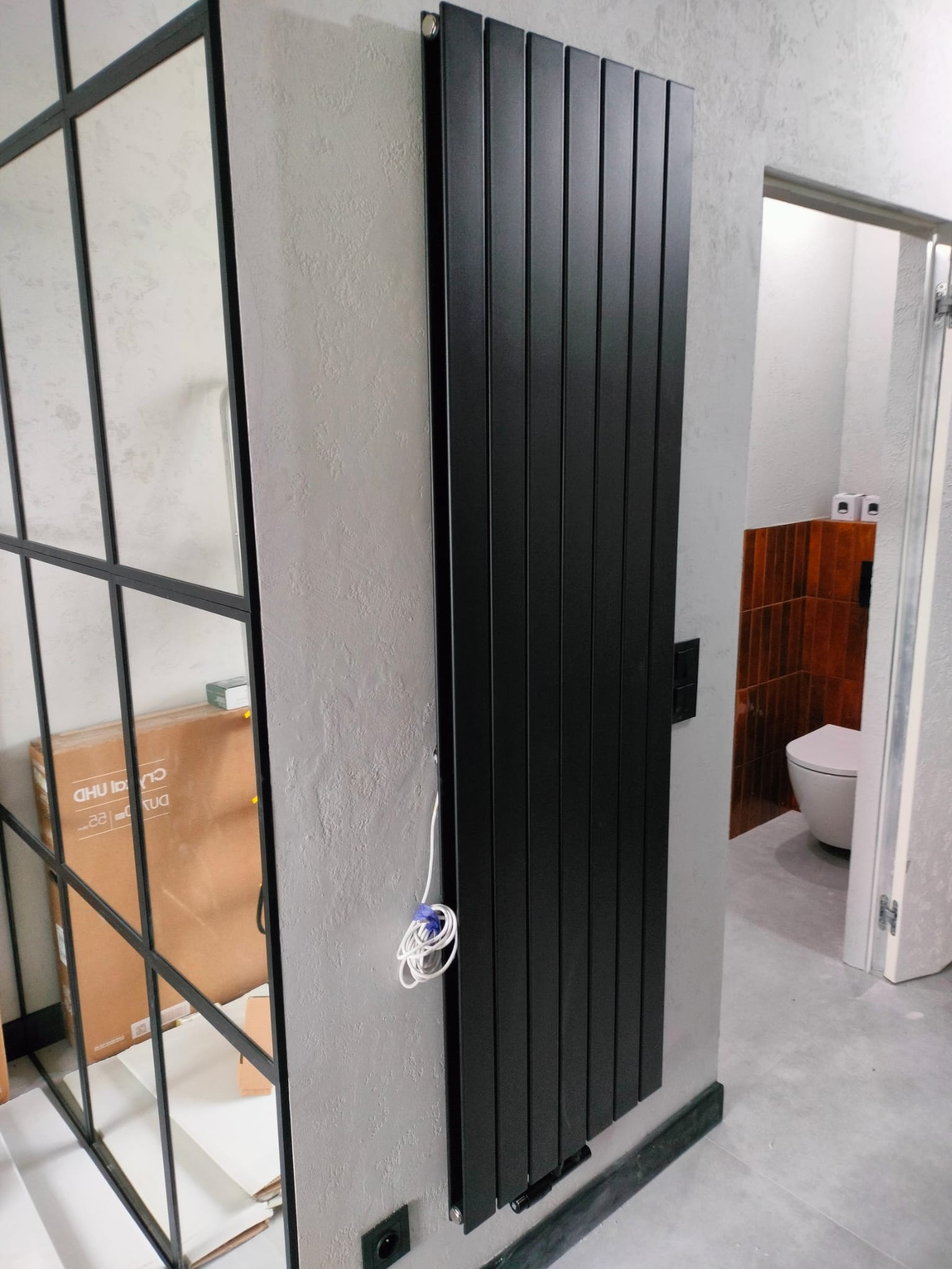 Czarny, pionowy grzejnik panelowy na szarej ścianie, widok z boku. W tle fragment łazienki z toaletą. Obok grzejnika widoczne przewody i skrzynka z szybami.