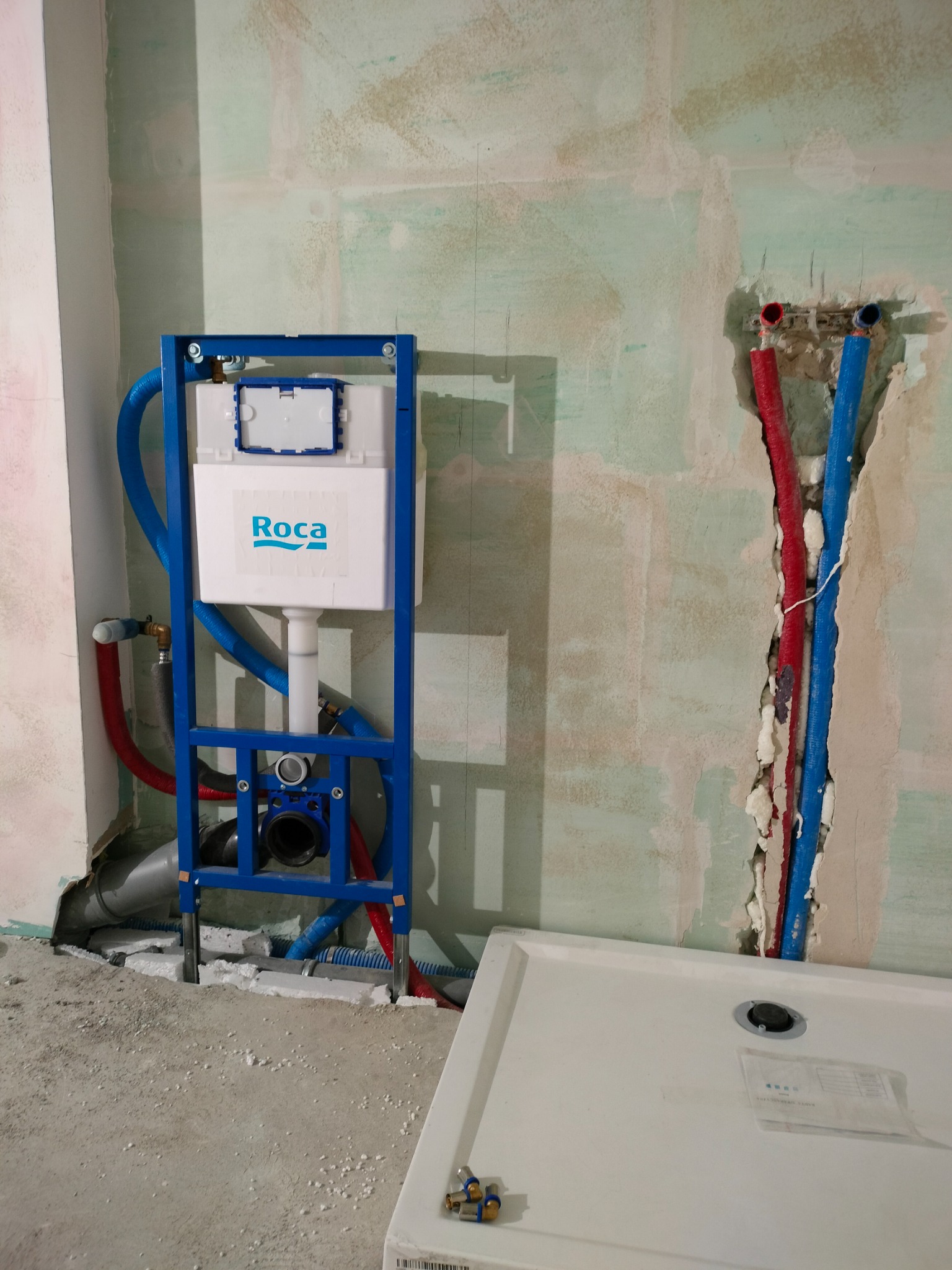 Stelaż WC Roca na tle surowej ściany z instalacją wodną (czerwone i niebieskie rury). Częściowo widoczny brodzik. Prace hydrauliczne w trakcie remontu łazienki.