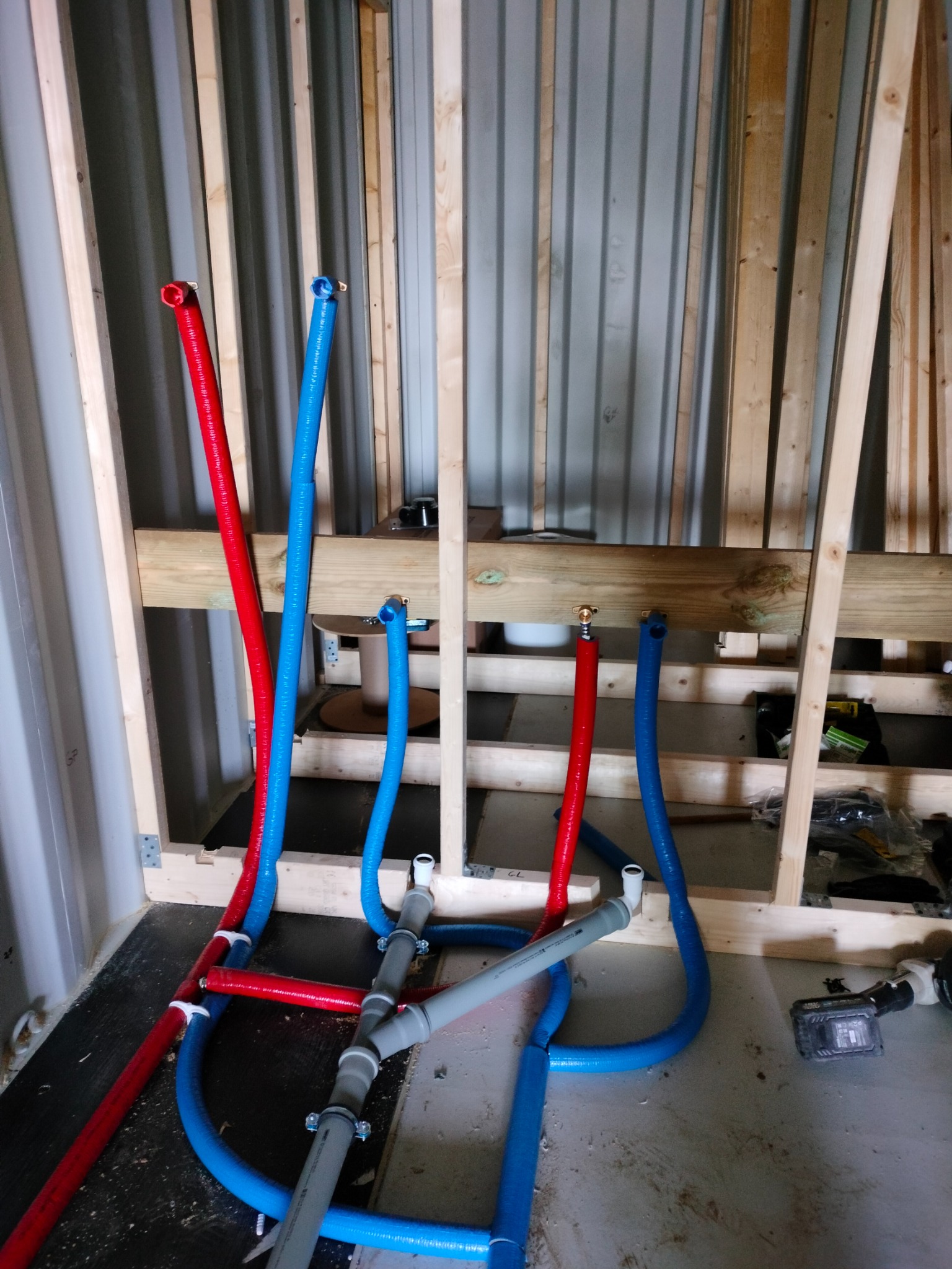 Instalacja wodna PEX: czerwone i niebieskie rury w drewnianej konstrukcji ściany z blachy falistej. Szare rury kanalizacyjne łączą się z kolorowymi.