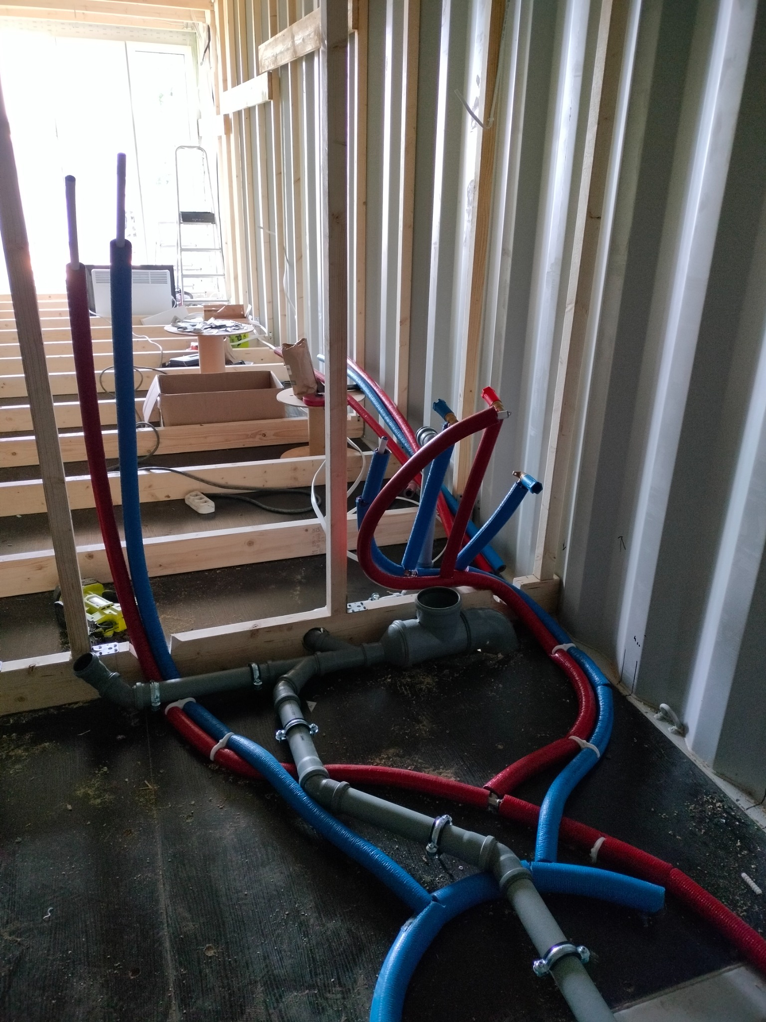 Instalacja wodna w trakcie montażu w surowym wnętrzu: rury PEX czerwone i niebieskie, szare rury kanalizacyjne, drewniana konstrukcja szkieletowa i blaszana ściana.
