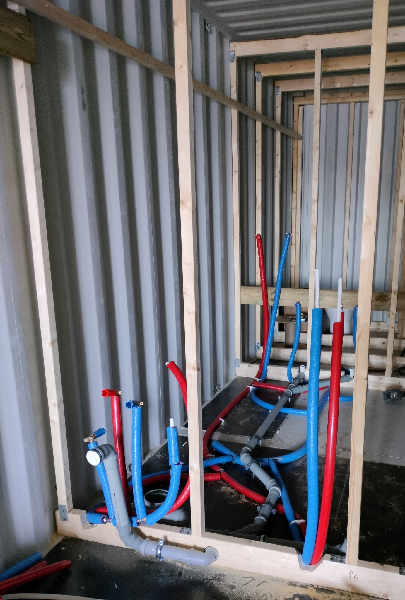 Instalacja wodna w kontenerze: czerwone i niebieskie rury, szare rury kanalizacyjne, drewniana konstrukcja szkieletowa, surowe ściany kontenera.