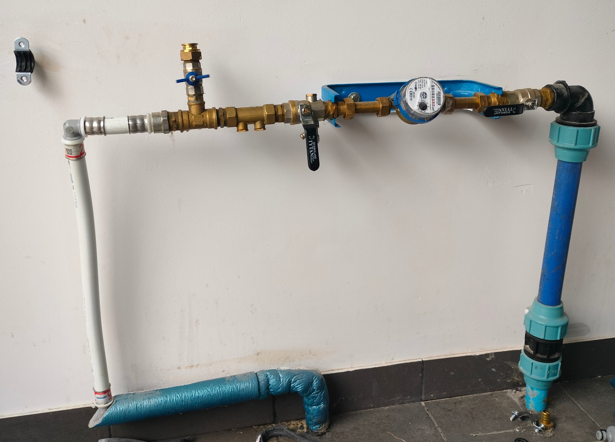 Kompletna instalacja wodna z licznikiem, zaworami i rurami z tworzywa sztucznego, zamontowana przy ścianie. Widoczne złącza, kolanka i izolacja termiczna.