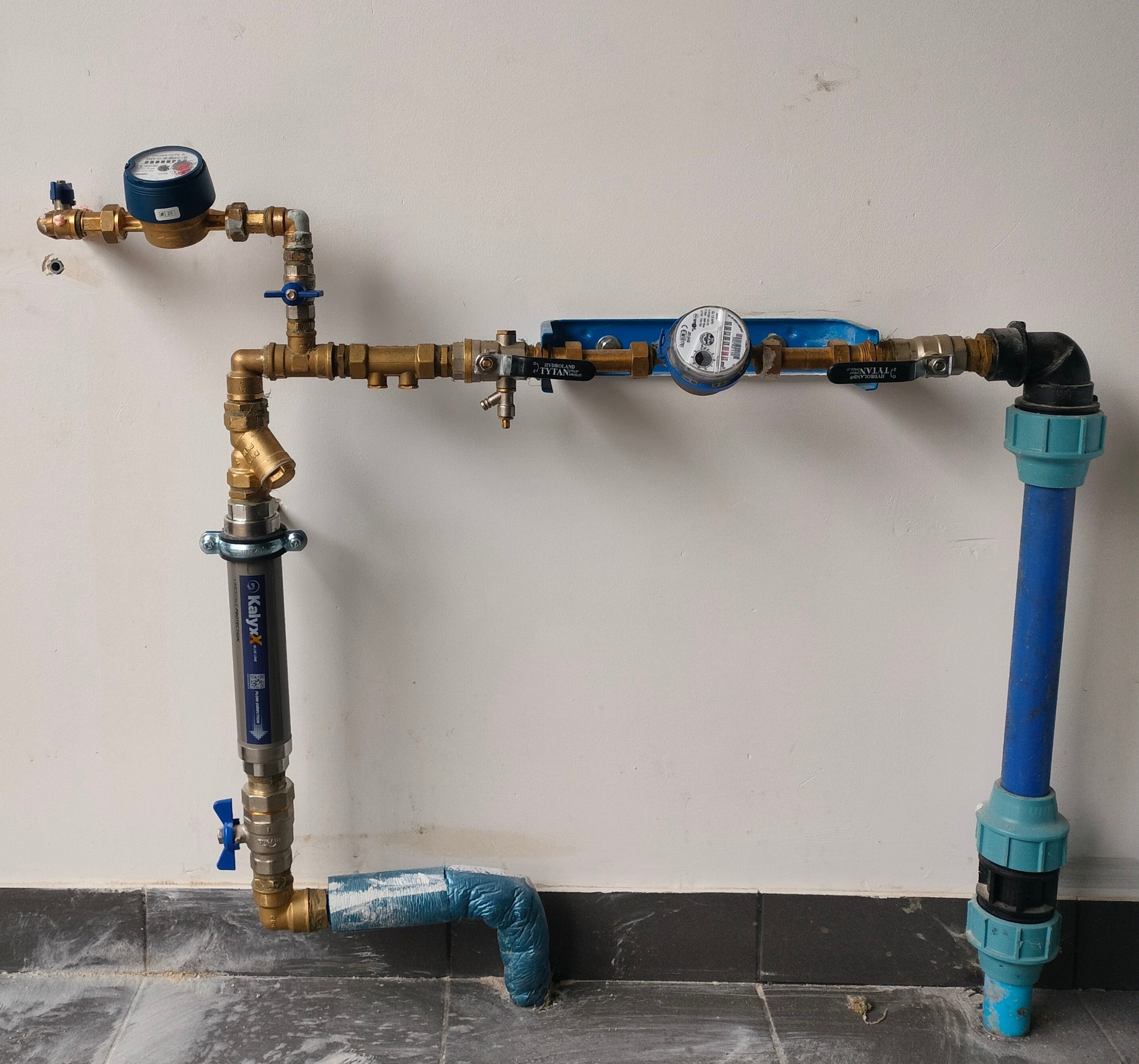 Kompletna instalacja wodna z filtrem i dwoma licznikami, wykonana z mosiądzu i niebieskiego PCV, zamontowana na białej ścianie z szarą listwą przypodłogową.