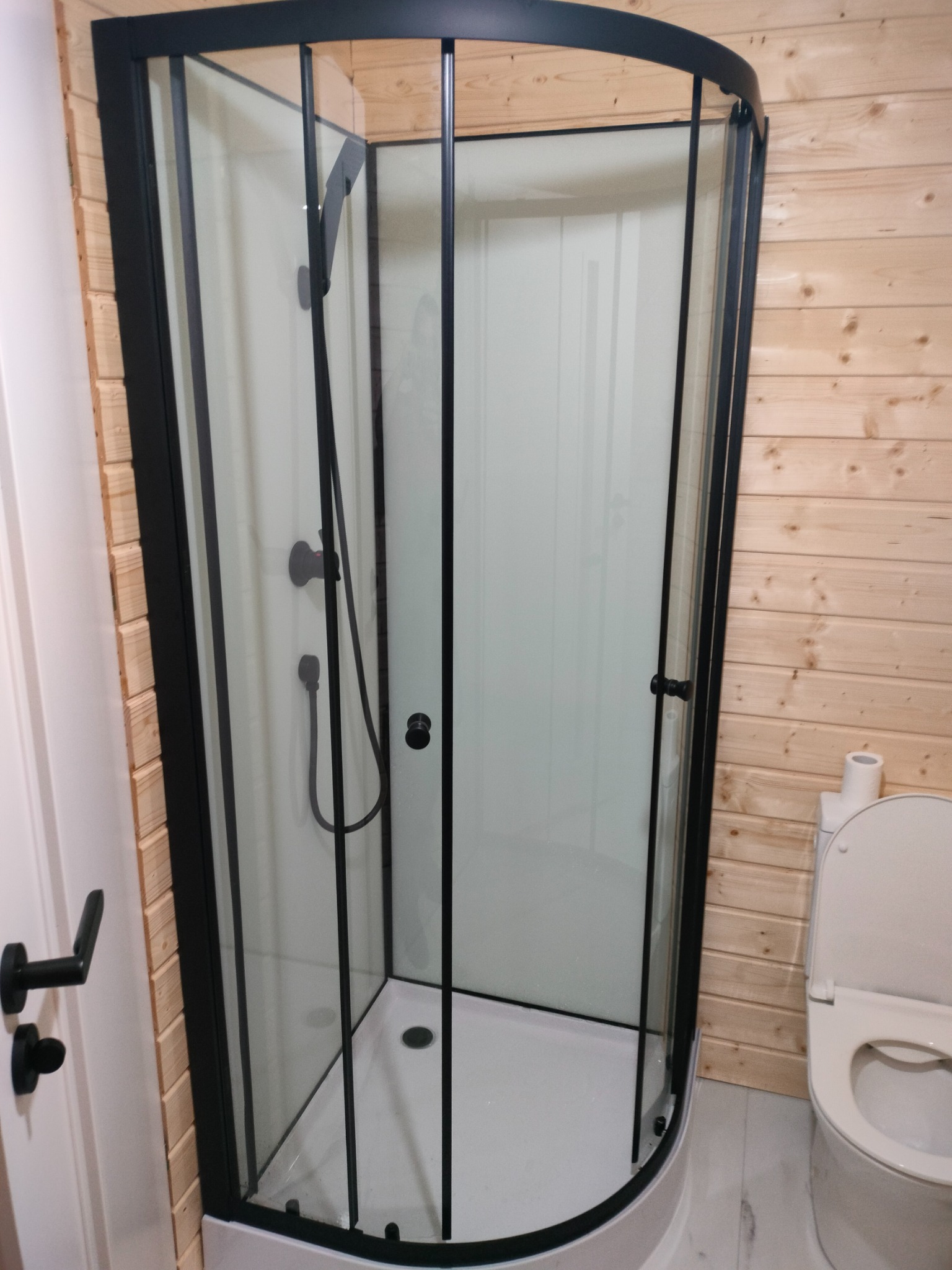 Kabina prysznicowa z czarnymi ramami w łazience wykończonej drewnem, widoczny fragment toalety i drzwi z czarną klamką. Minimalistyczny design.