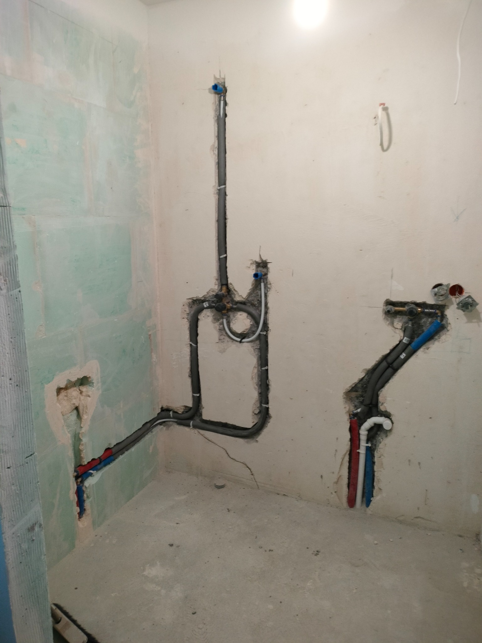 Surowe ściany z instalacją hydrauliczną w trakcie montażu: rury szare, niebieskie i czerwone, widoczne punkty podłączeń. Etap prac przed wykończeniem.