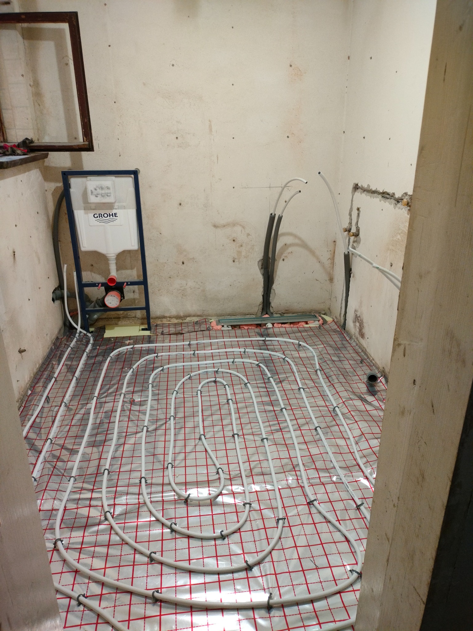Białe rury ogrzewania podłogowego ułożone na srebrnej folii z czerwoną siatką w remontowanej łazience. Widoczny stelaż podtynkowy Grohe.
