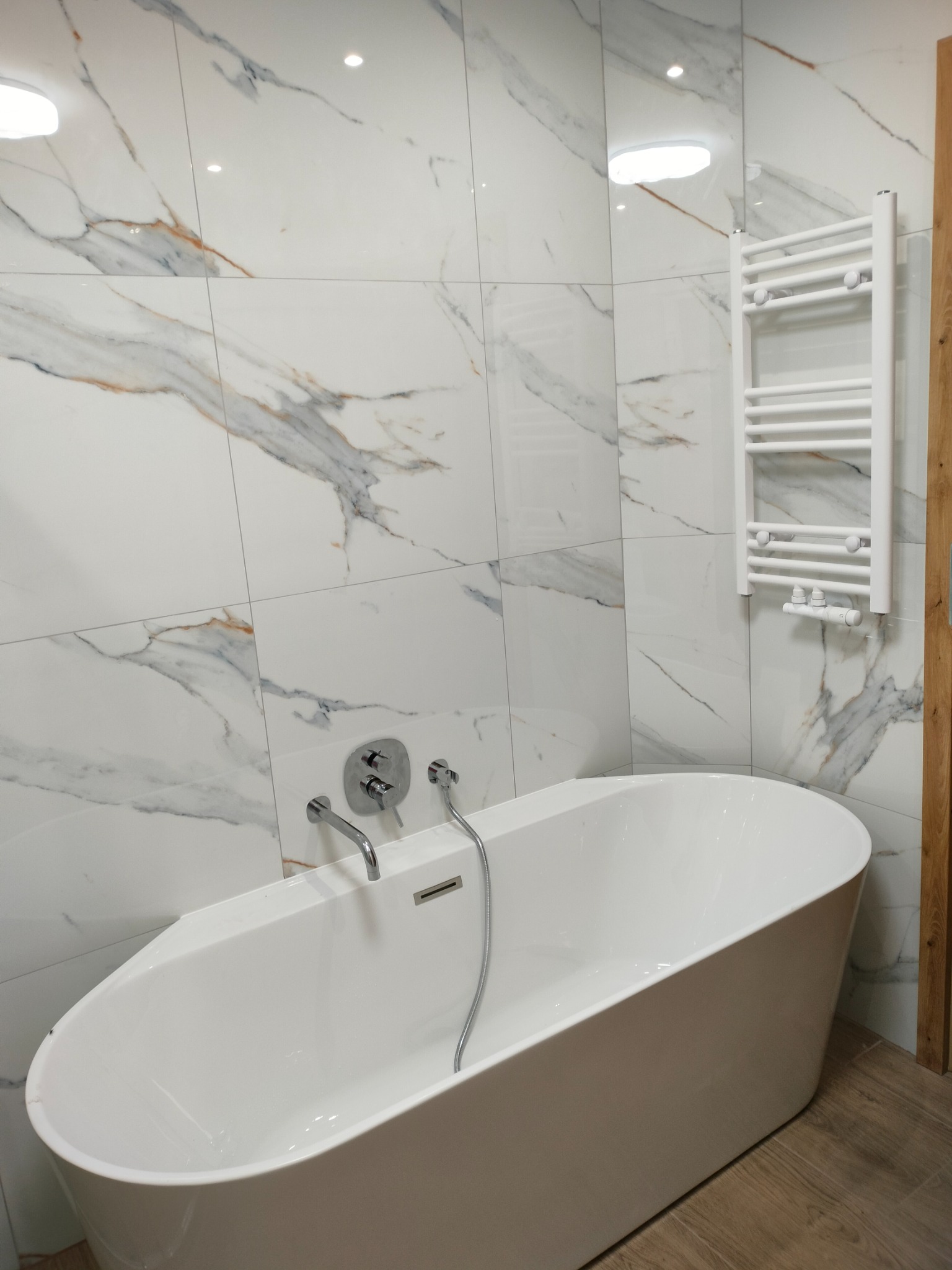 Biała, wolnostojąca wanna z chromowaną armaturą na tle marmurowych płytek i grzejnika drabinkowego w nowoczesnej łazience.