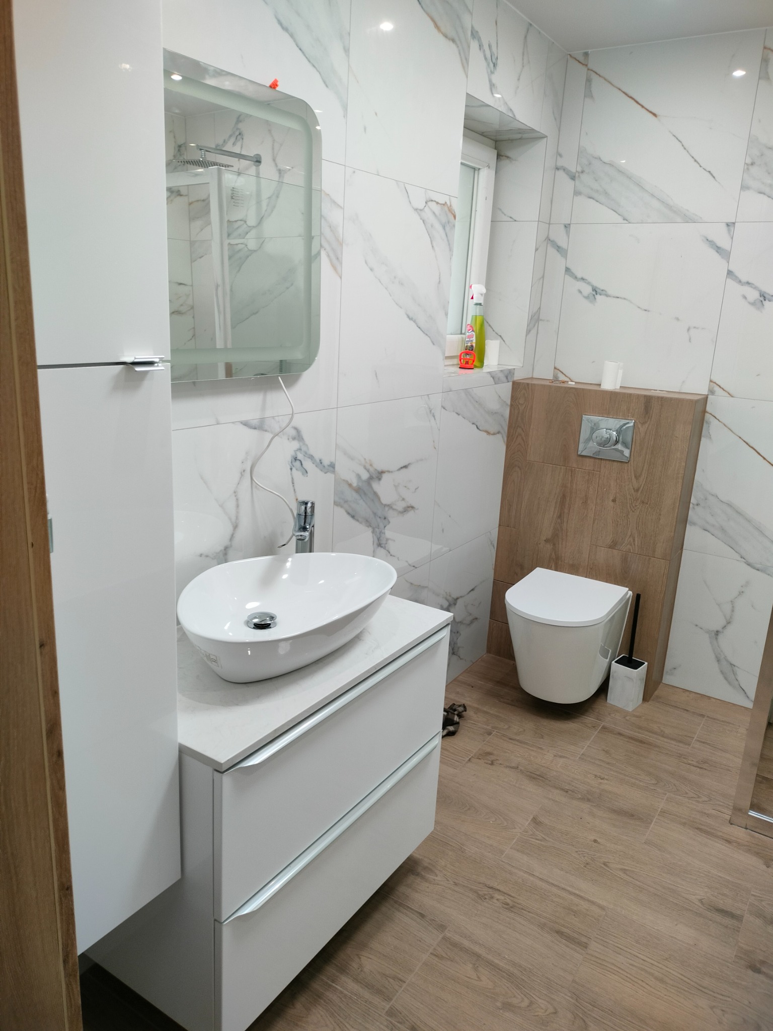 Nowoczesna łazienka z białą umywalką nablatową, podwieszaną toaletą na drewnianym panelu i marmurowymi płytkami. Minimalistyczny design, jasne wnętrze.