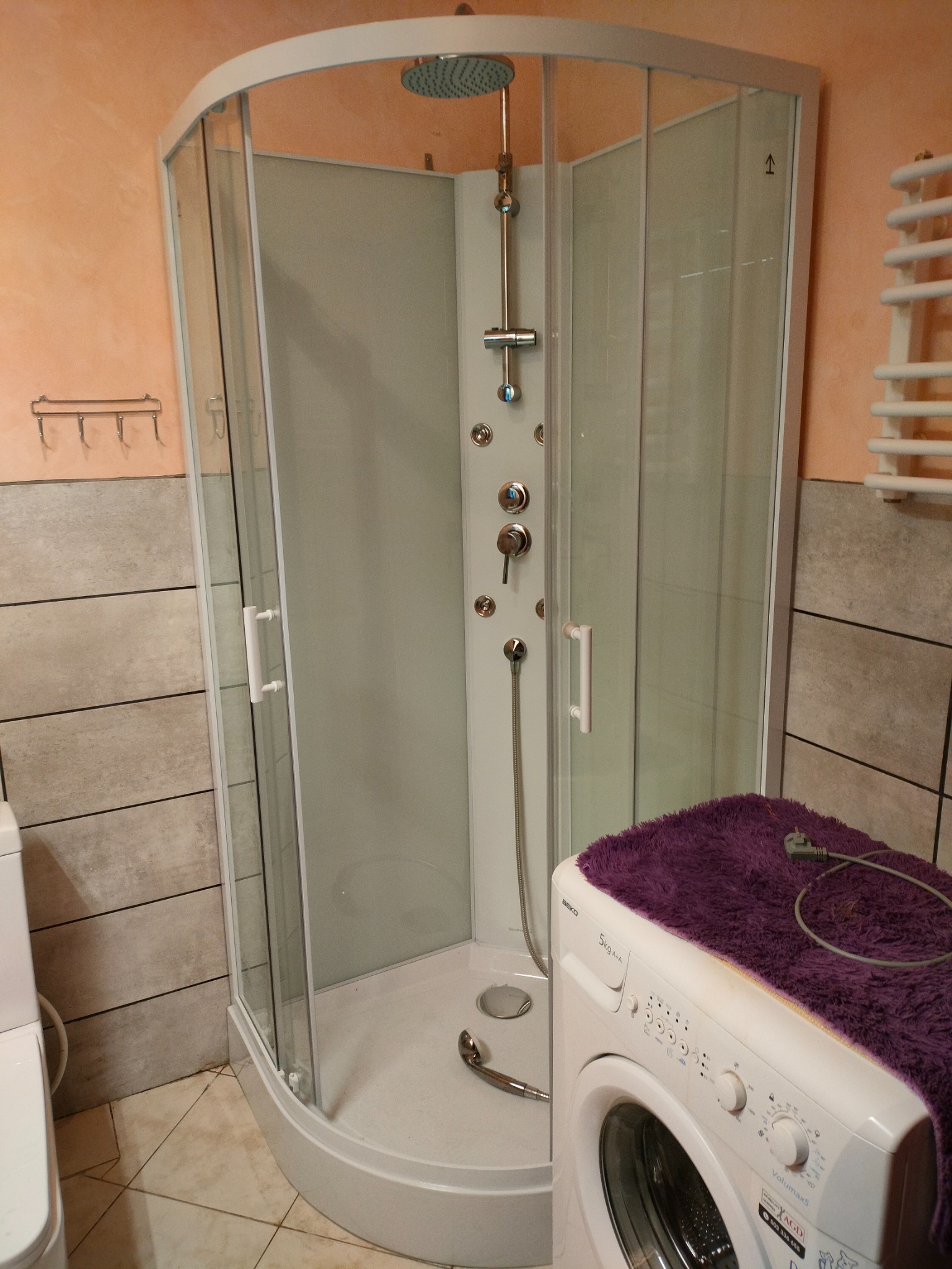 Kabina prysznicowa z hydromasażem w łazience, obok pralka z fioletowym ręcznikiem. Widoczne płytki na ścianach i grzejnik drabinkowy.
