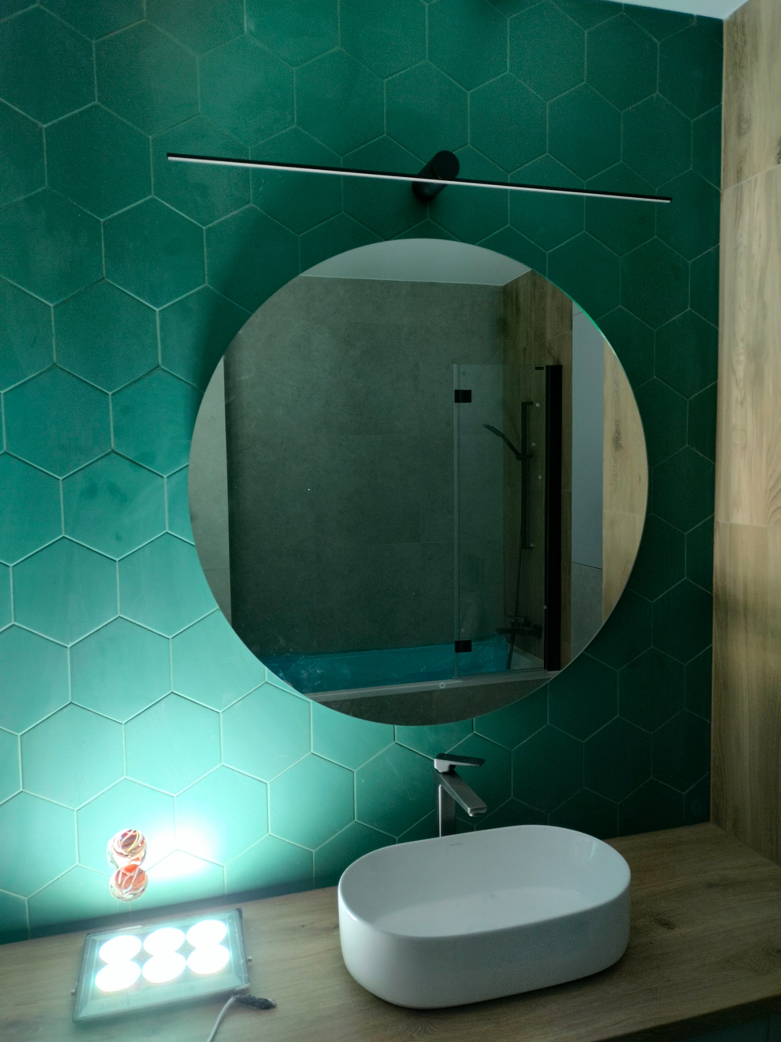 Nowoczesna łazienka z umywalką nablatową i okrągłym lustrem na tle ściany wyłożonej zielonymi, heksagonalnymi płytkami. Widoczna bateria i oświetlenie.