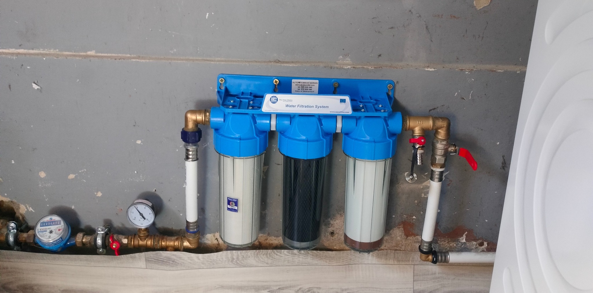 Niebieski system filtracji wody, z trzema przezroczystymi pojemnikami, podłączony do rur miedzianych i białych plastikowych, zamontowany na szarej ścianie.