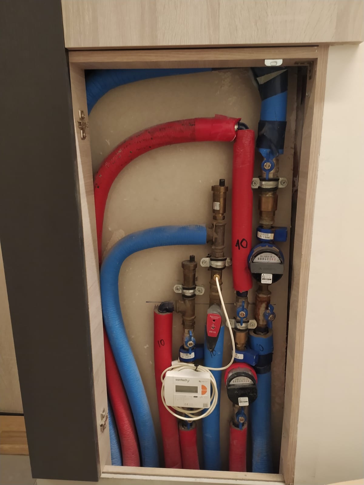 Węzeł wodny z rurami w izolacji (czerwone, niebieskie) i licznikiem. Instalacja hydrauliczna w szafce w Orlu. Widoczne zawory i elementy przyłączeniowe.