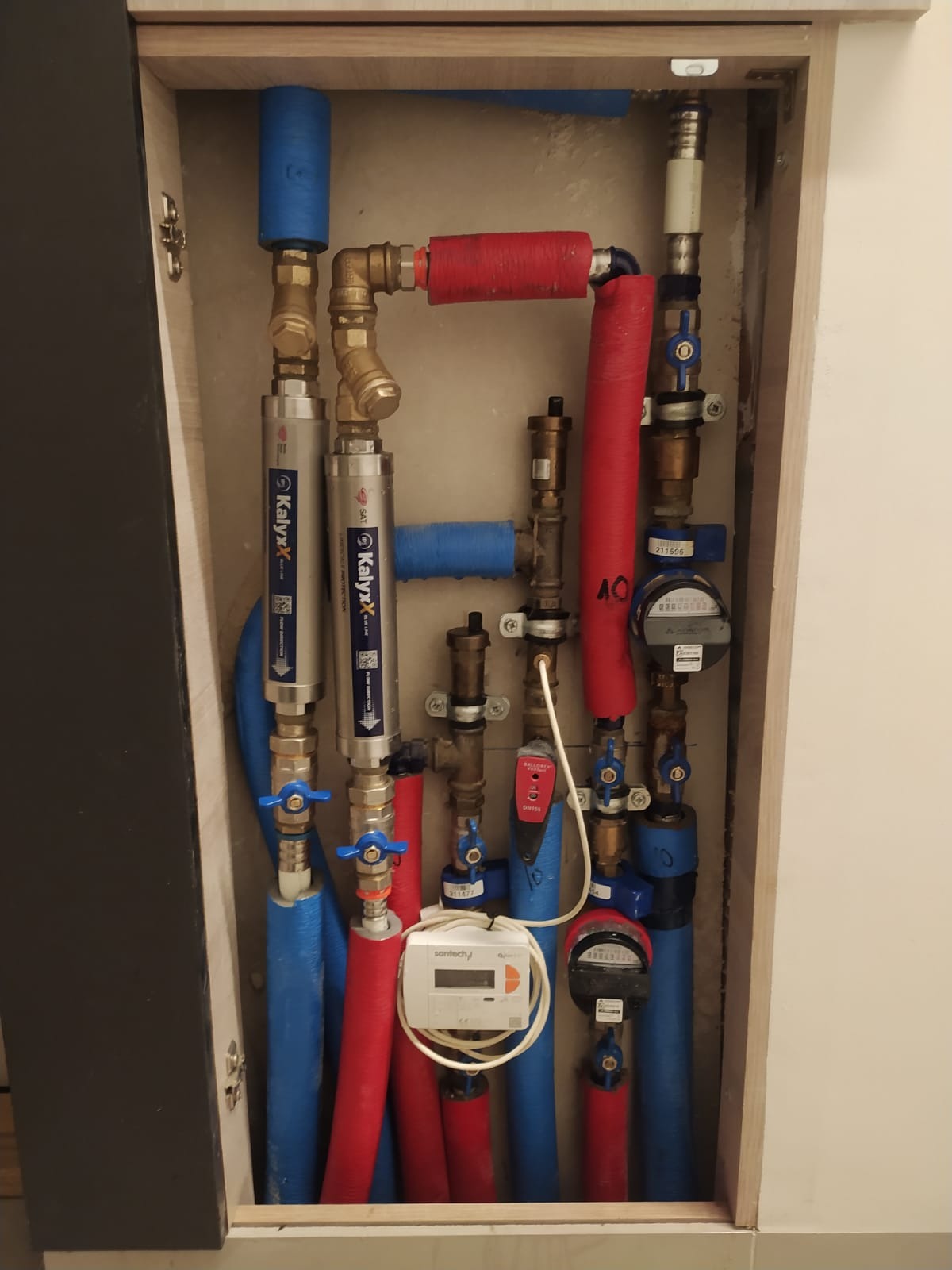 Węzeł cieplny w szafce: rury w izolacji czerwonej i niebieskiej, zawory, filtry marki Kalyxx i liczniki. Widoczne elementy instalacji wodnej i grzewczej.