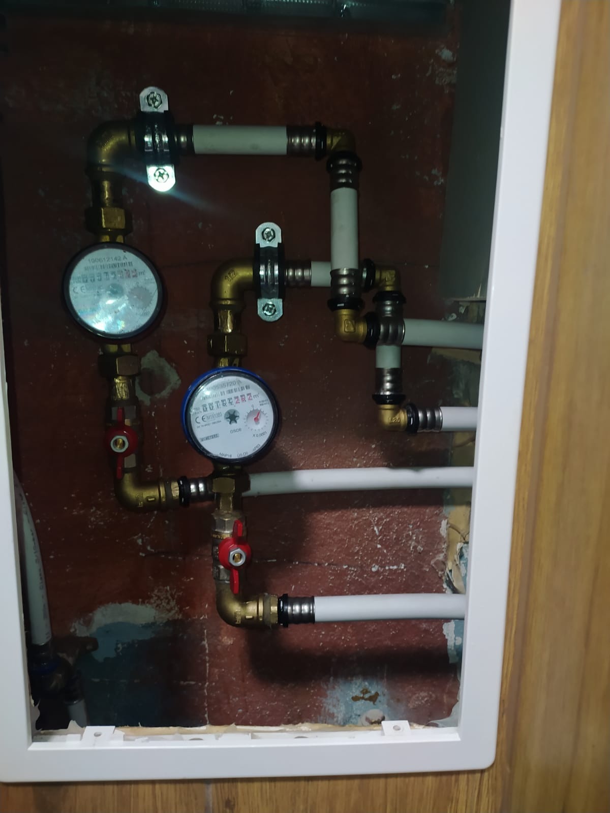 Złożona instalacja wodna z dwoma licznikami, mosiężnymi złączkami i białymi rurami w obudowie, widoczne zawory odcinające i elementy mocujące.