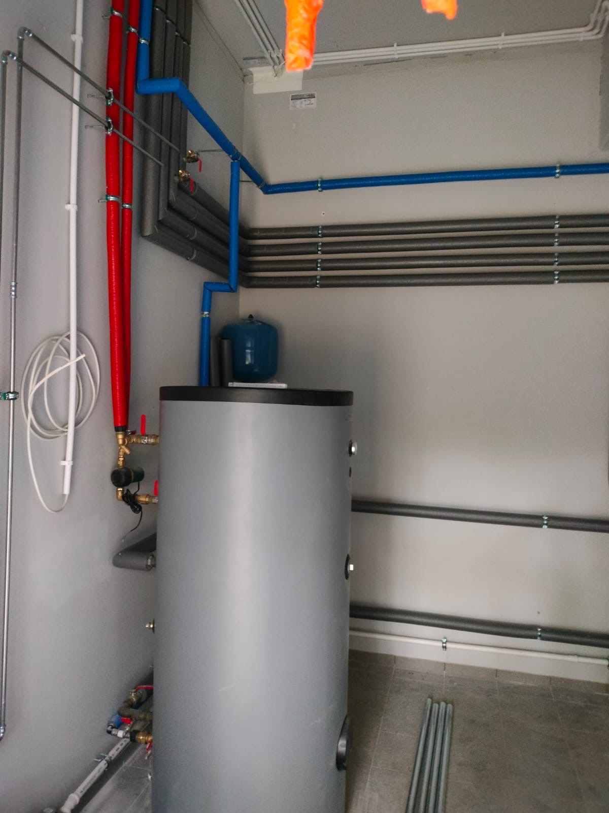 Szary bojler z niebieskim zbiornikiem wyrównawczym, otoczony instalacją z rur w kolorach: czerwonym, niebieskim i szarym. Widoczne mocowania rur do ściany.