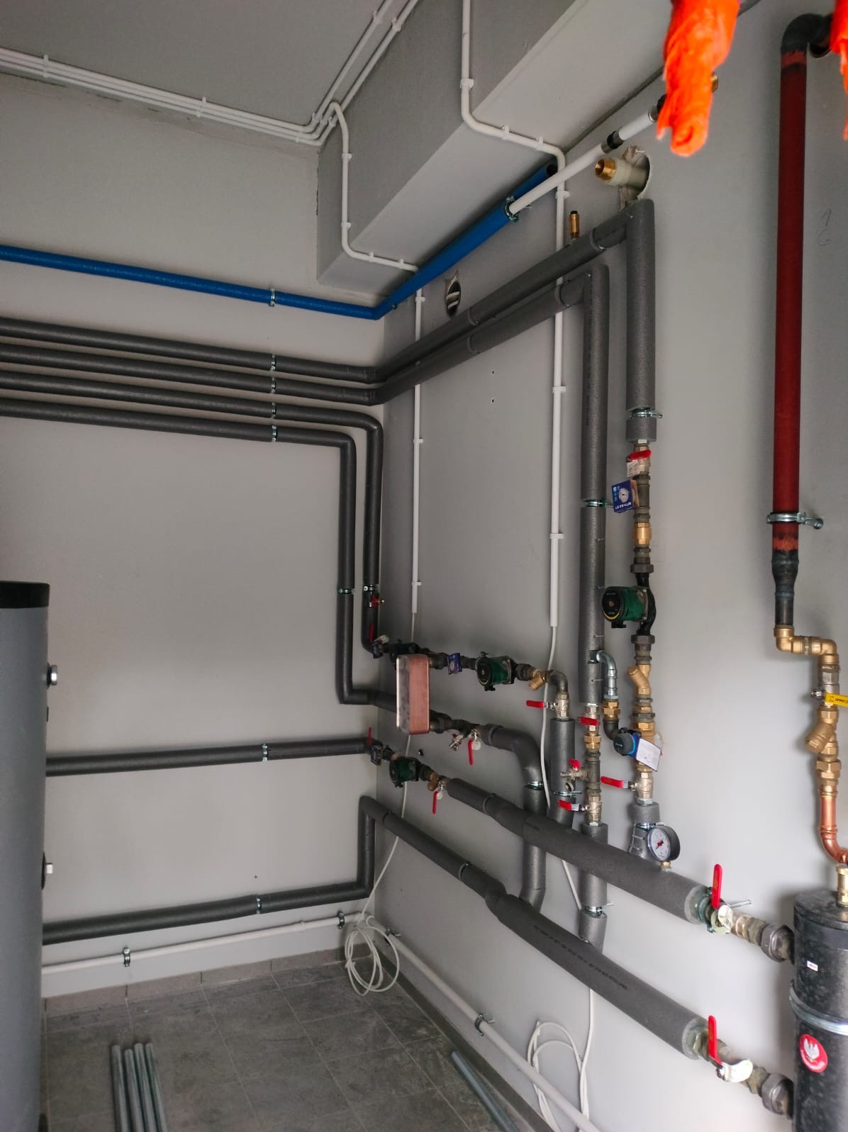 Szare rury grzewcze z izolacją, zawory i manometry na ścianie. Instalacja CO w kotłowni, widoczne elementy instalacji hydraulicznej i grzewczej.