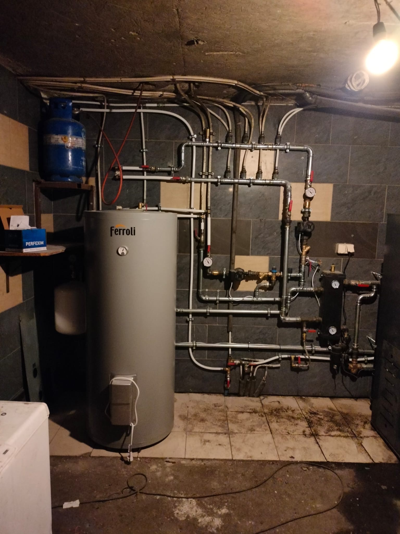 Szary piec Ferroli w kotłowni z widoczną instalacją hydrauliczną na ścianie z szarych płytek. Na półce butla gazowa i pudełko Perfexim.