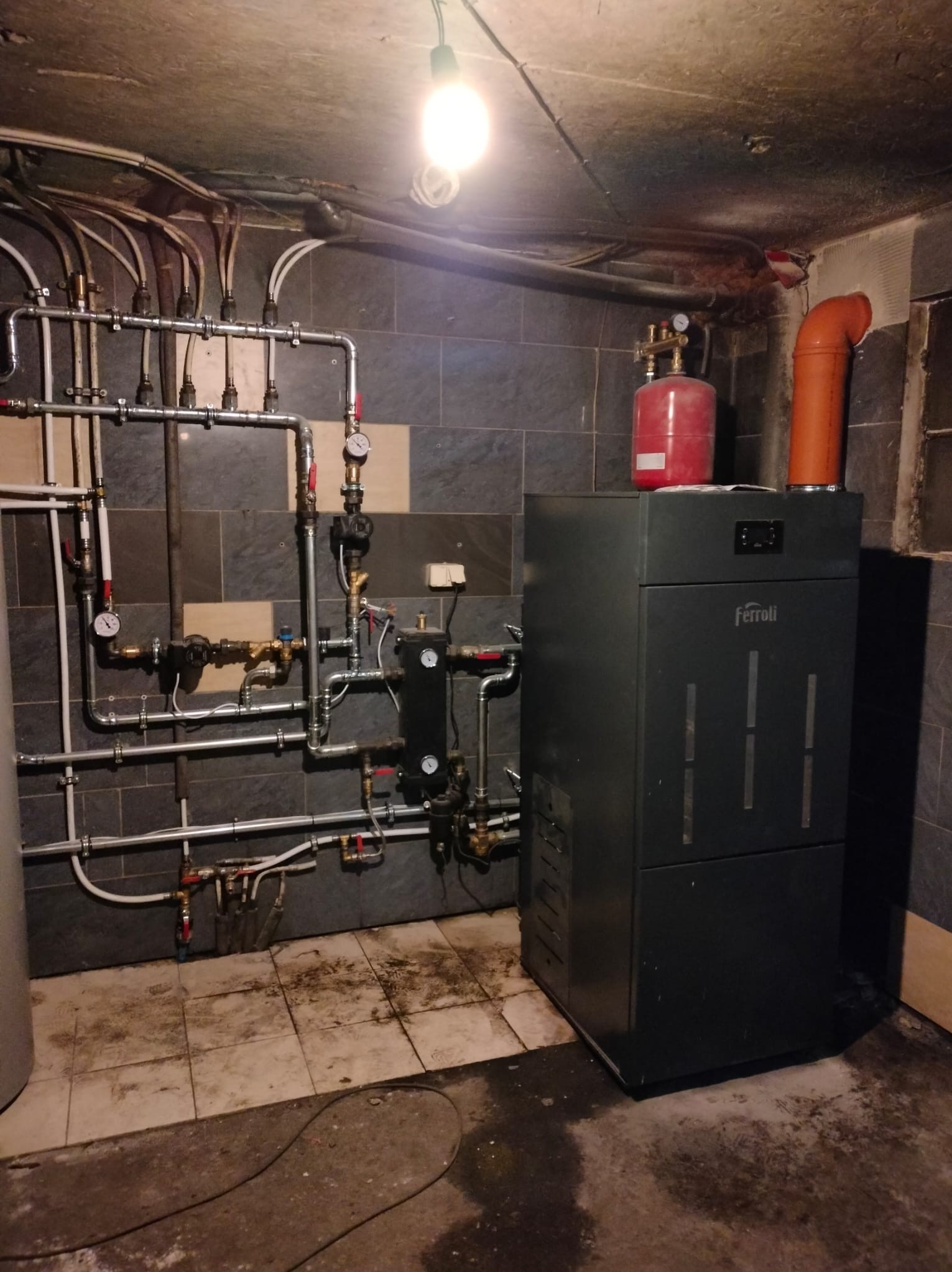 Czarny piec Ferroli z instalacją grzewczą w kotłowni z szarymi płytkami. Widoczne rury, zawory i manometry. Oświetlenie górne.