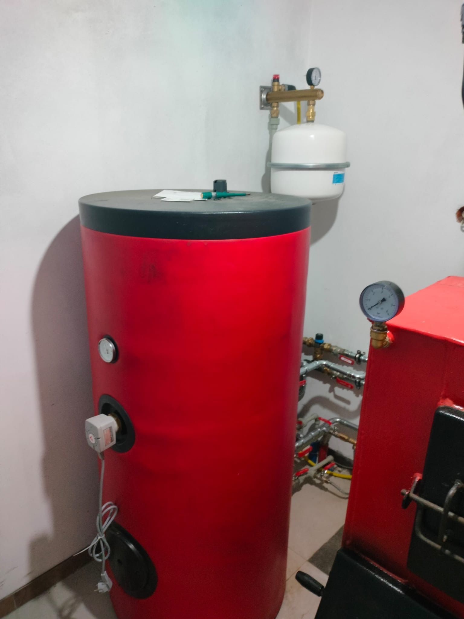 Czerwony bojler z czarnym blatem w kotłowni, obok piec CO z manometrem. Widoczne rury instalacji hydraulicznej i zawory. Białe ściany i jasna podłoga.