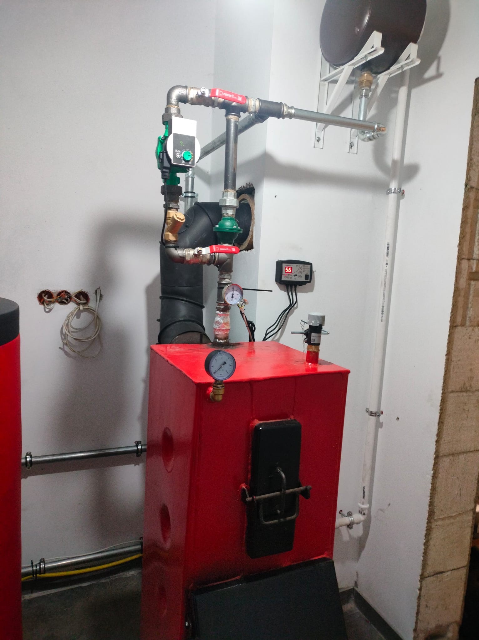 Czerwony piec CO z czarnymi drzwiczkami i manometrem, podłączony do instalacji z zaworami i sterownikiem na ścianie. Widoczne rury i przewody.