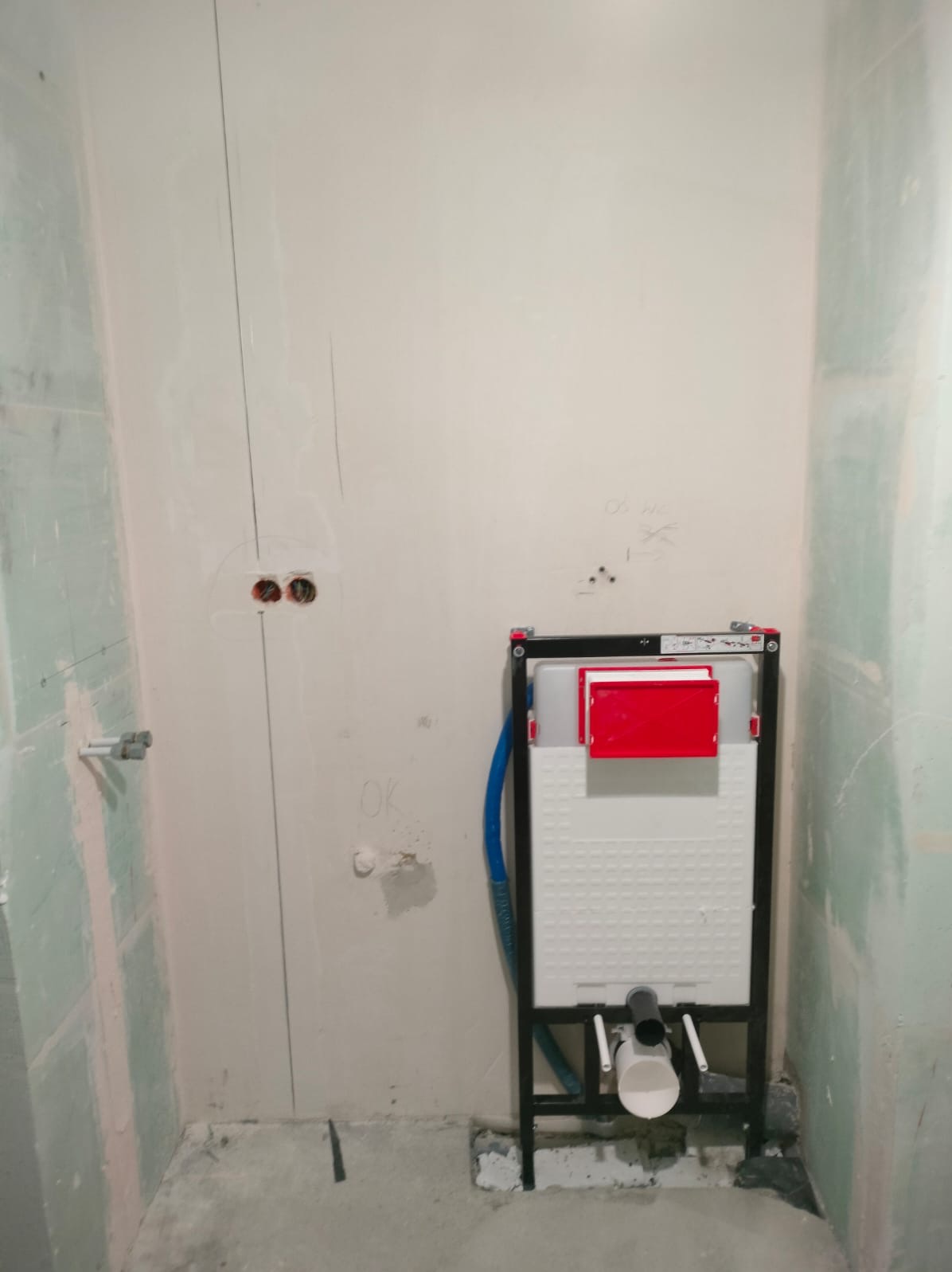 Stelaż podtynkowy WC z czerwoną klapką, zamontowany na surowej ścianie w trakcie prac wykończeniowych. Widoczne instalacje i ślady po wierceniu.