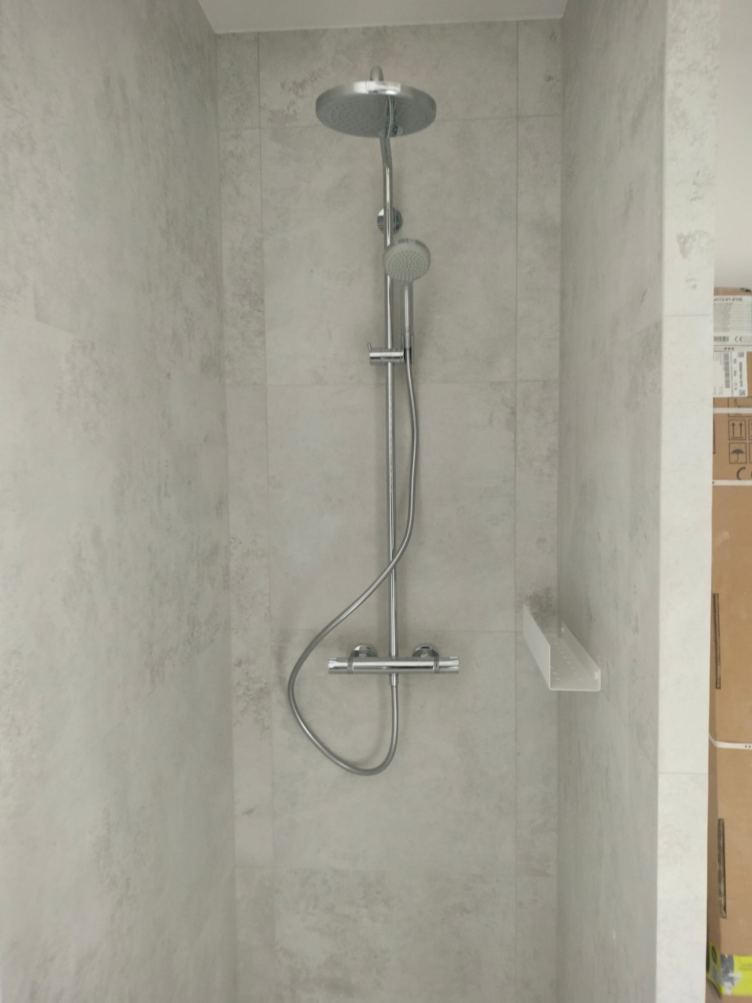 Nowoczesny zestaw prysznicowy z deszczownicą i termostatem, zamontowany na ścianie wyłożonej jasnymi, marmurkowymi płytkami. Minimalistyczna półka w rogu kabiny.