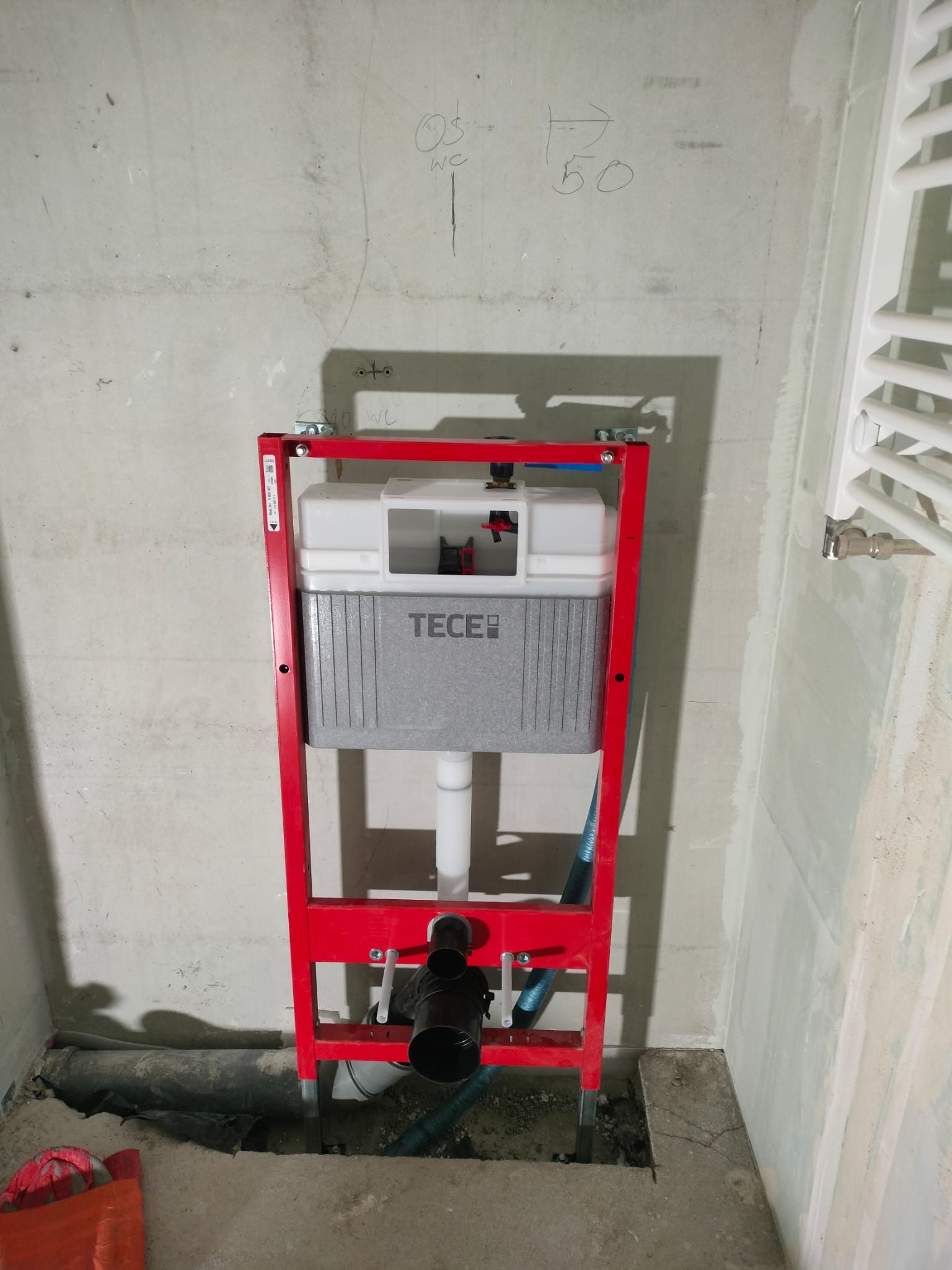 Stelaż podtynkowy WC marki TECE w czerwonej ramie, zamontowany w surowym wnętrzu łazienki z widocznymi instalacjami i grzejnikiem drabinkowym.