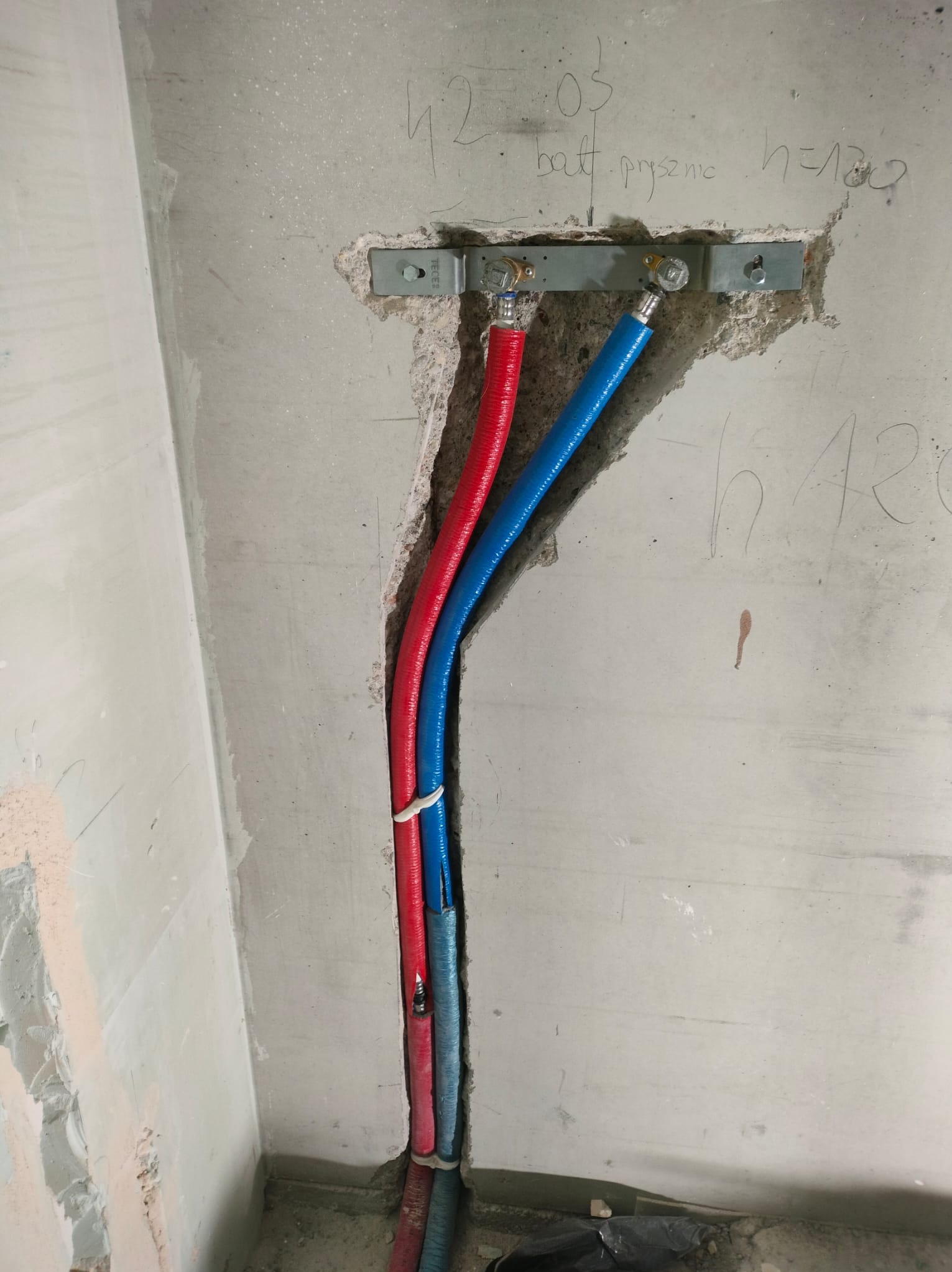 Czerwona i niebieska rura wodna zamontowana w surowej ścianie z betonu, zabezpieczona metalową listwą. Widoczne napisy na ścianie i ślady po kuciu.