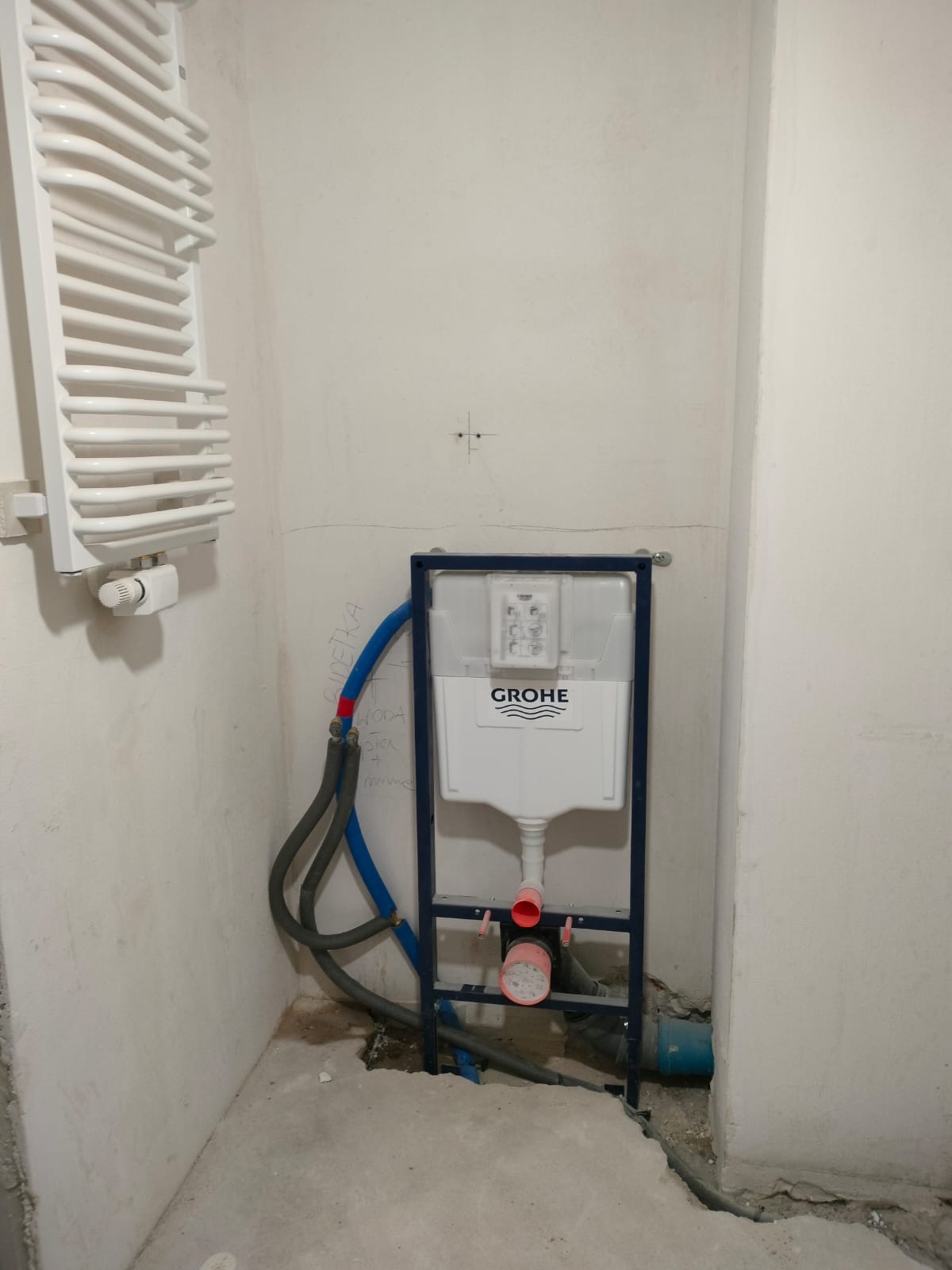 Stelaż podtynkowy Grohe z podłączonymi rurami w surowej łazience, obok grzejnik drabinkowy. Widoczne ślady po instalacji na ścianie.