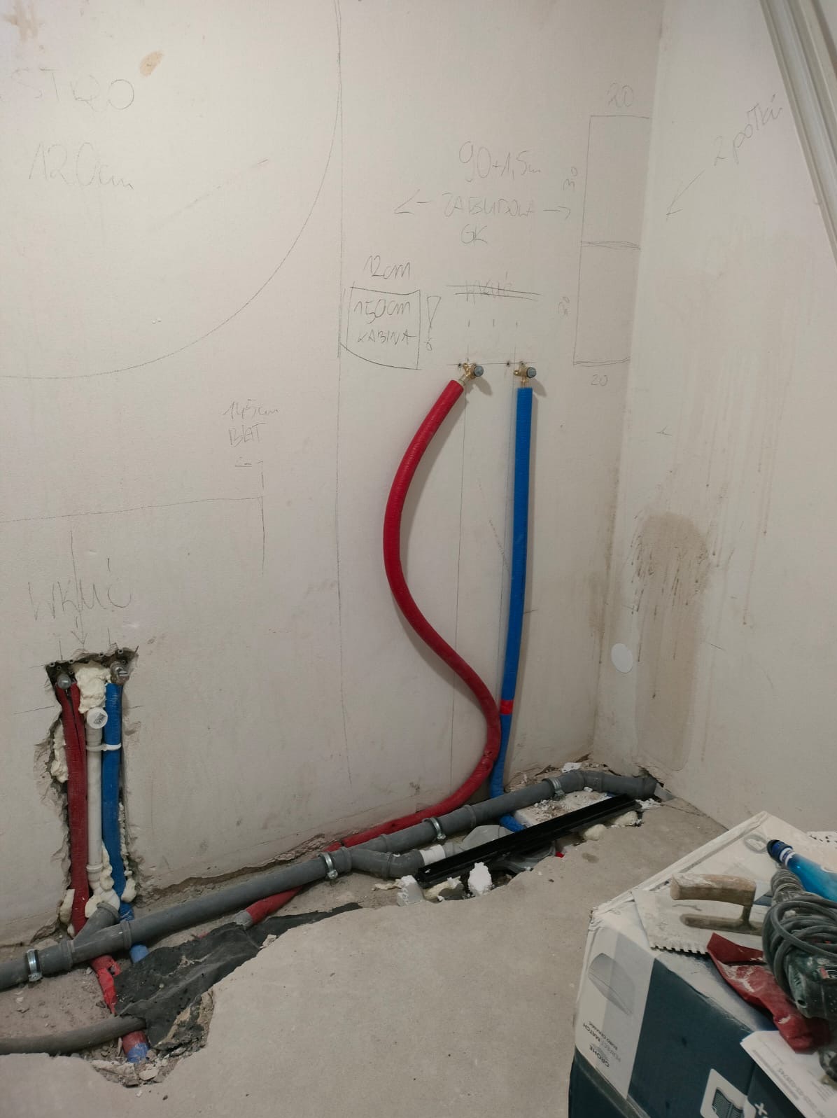 Instalacja wodna w łazience w trakcie remontu. Widoczne rury w kolorze czerwonym i niebieskim, szare rury kanalizacyjne oraz notatki na ścianie dotyczące wymiarów.