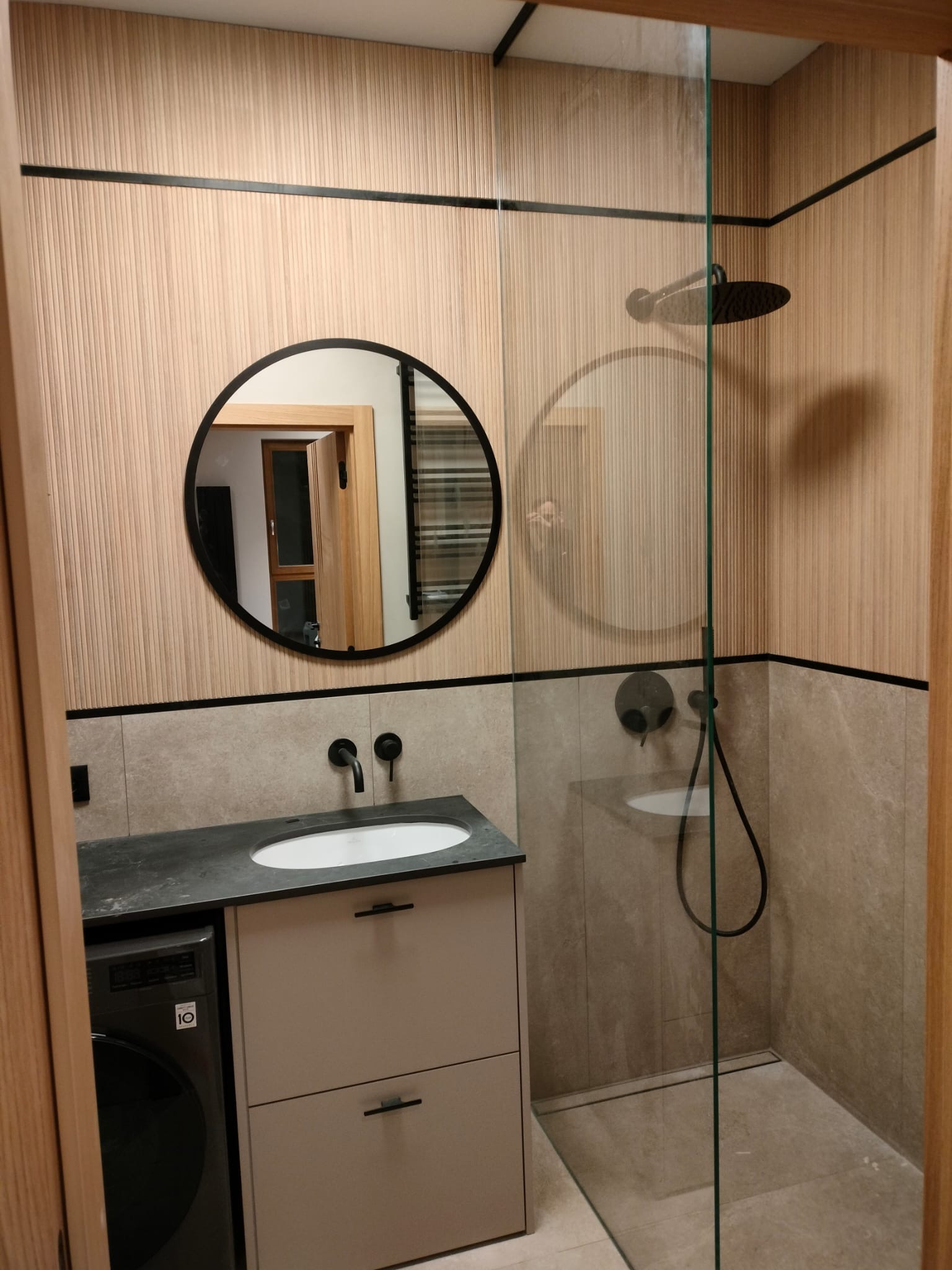 Nowoczesna łazienka z kabiną prysznicową typu walk-in, umywalką nablatową i okrągłym lustrem. Ściany wykończone drewnopodobnymi panelami i szarą płytką.