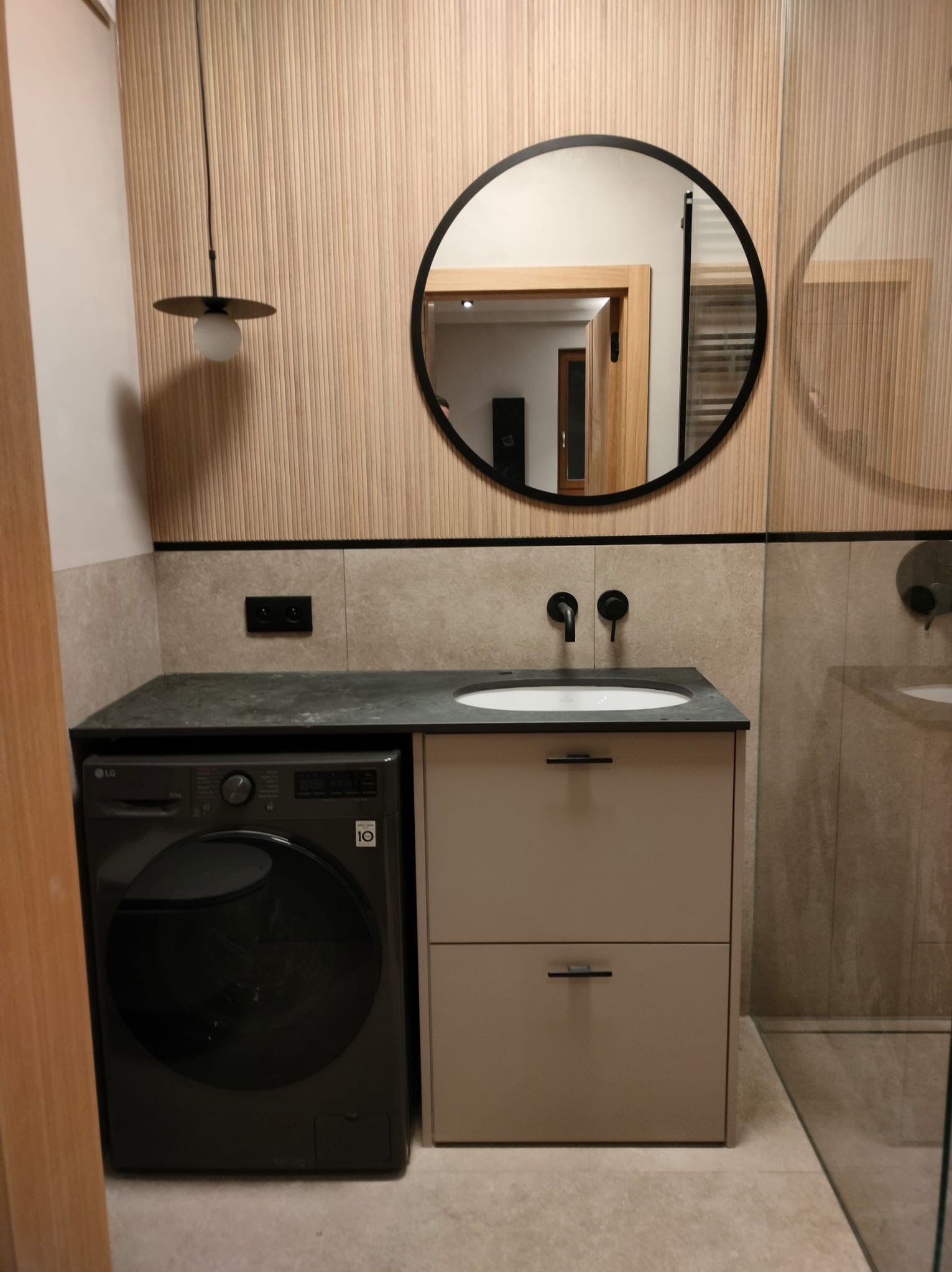 Nowoczesna łazienka z pralką LG obok szafki umywalkowej z czarną armaturą, okrągłe lustro i dekoracyjne panele ścienne. Minimalistyczny design.