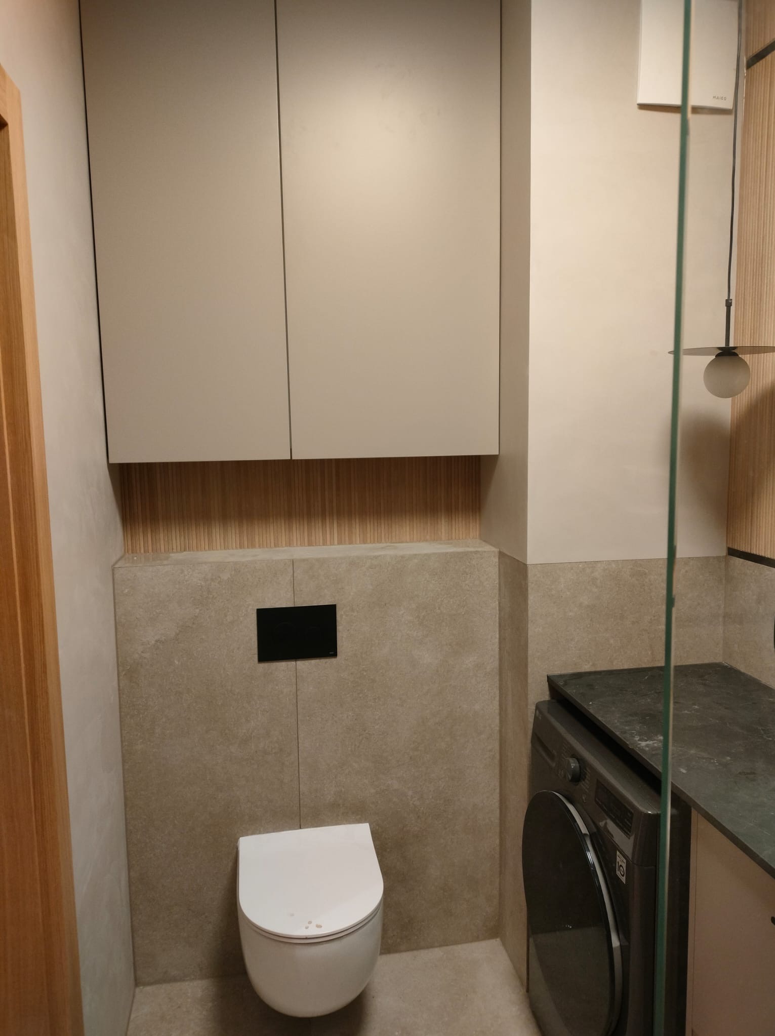 Nowoczesna łazienka w beżach: podwieszana toaleta z czarnym przyciskiem, szafki nad nią, pralka pod blatem i szklana kabina prysznicowa. Minimalistyczny design.