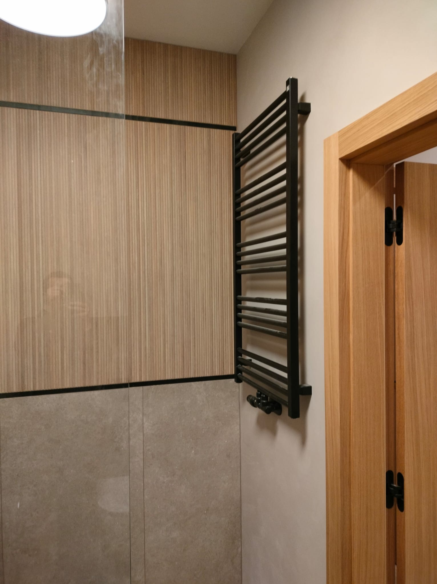 Czarny, drabinkowy grzejnik łazienkowy na ścianie z drewnianą okładziną i szarą płytką, obok drewniane drzwi z czarnymi zawiasami. Nowoczesne wykończenie łazienki.
