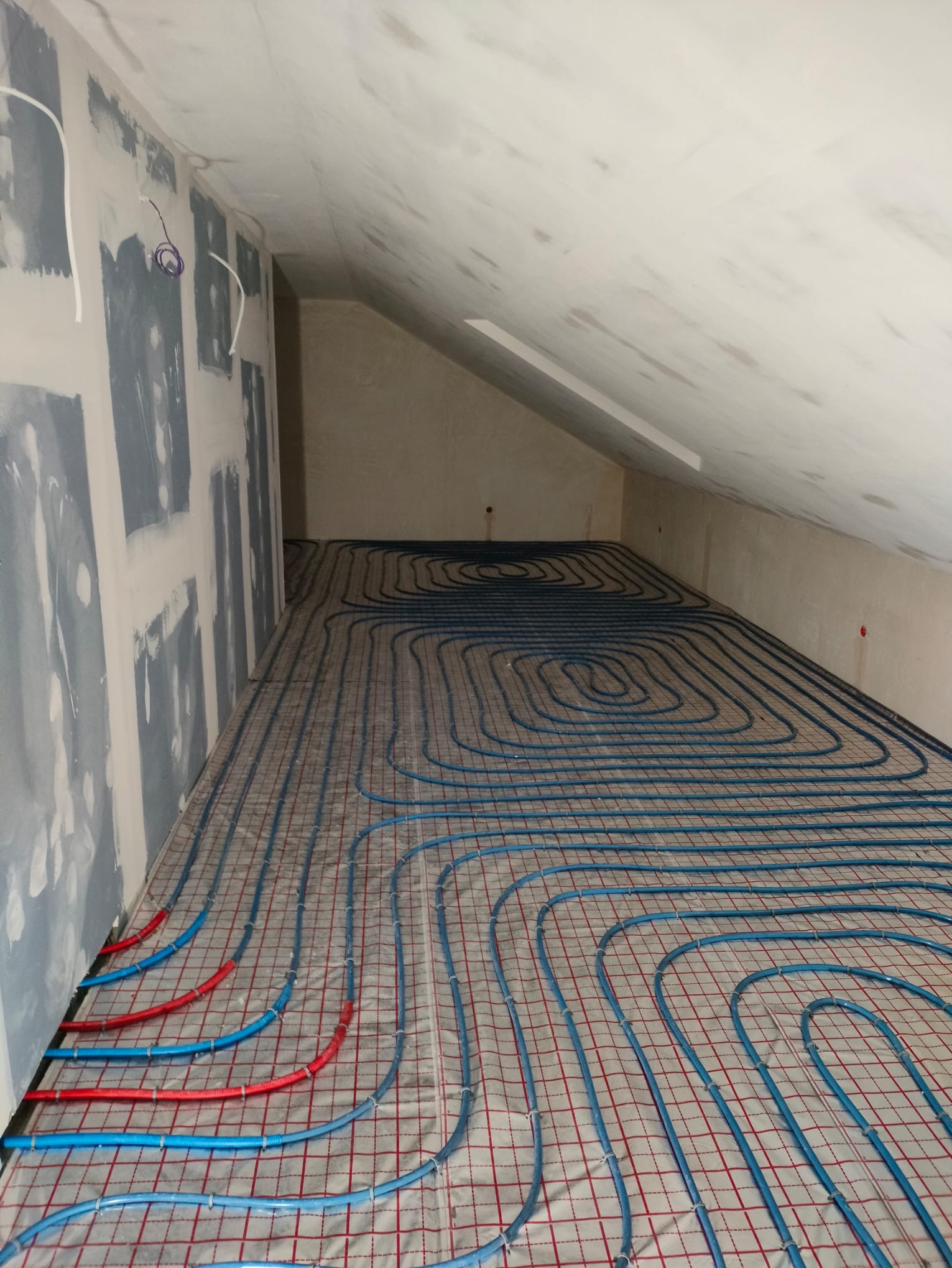 Niebieskie rury ogrzewania podłogowego ułożone na siatce w surowym wnętrzu poddasza, widoczne fragmenty ścian przygotowanych do wykończenia.