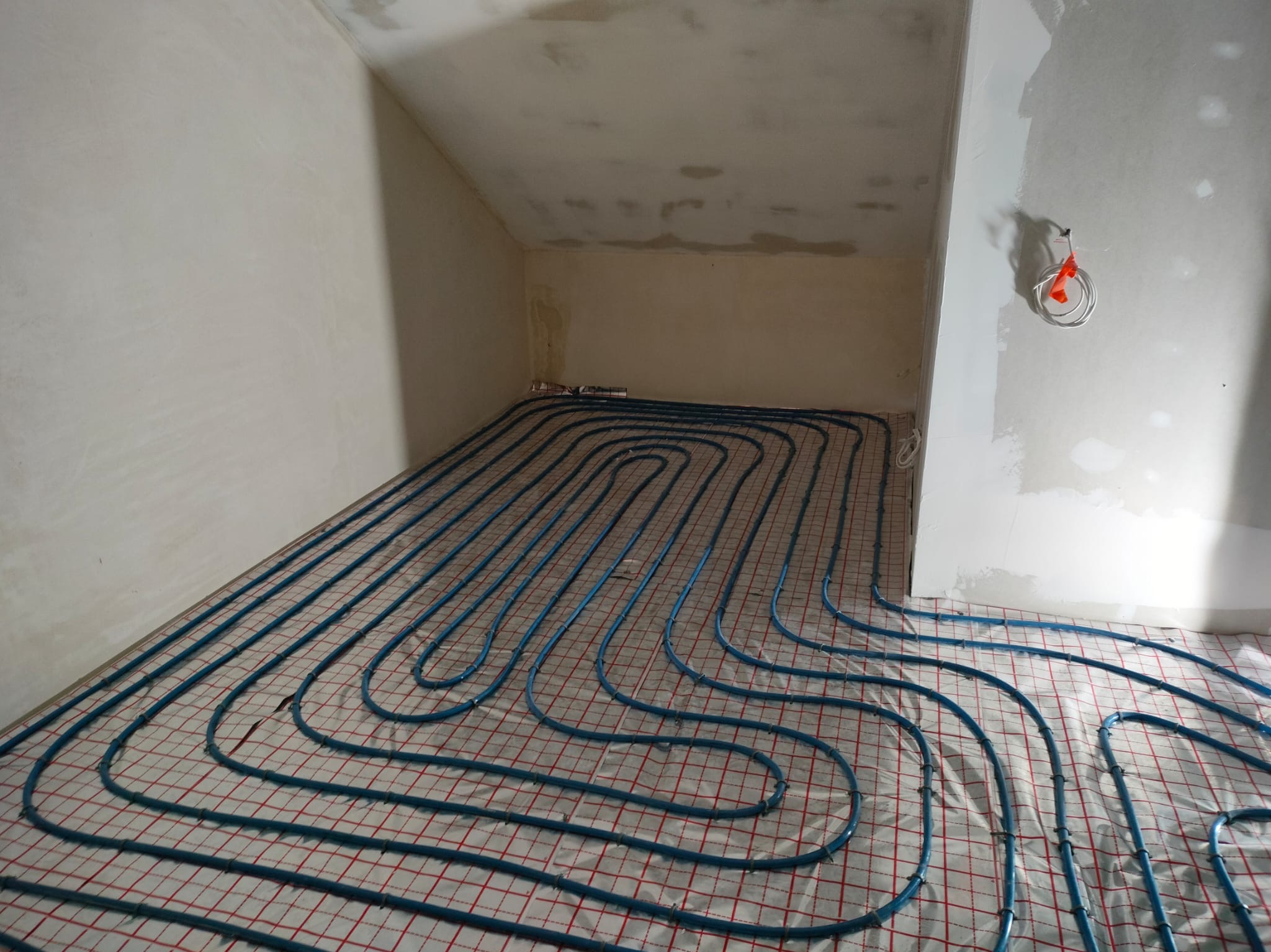 Niebieskie rury ogrzewania podłogowego ułożone na siatce w surowym wnętrzu. Widoczne niedokończone ściany i instalacja elektryczna.