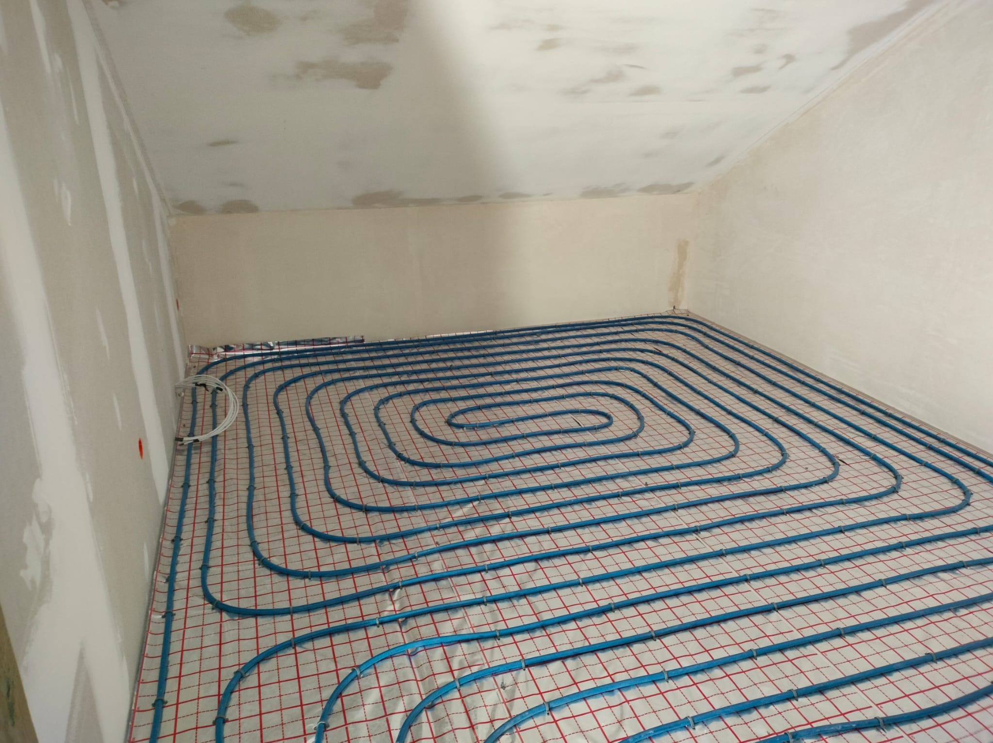 Niebieskie rury ogrzewania podłogowego ułożone spiralnie na izolacji z czerwoną siatką w surowym wnętrzu. Widok z góry na instalację przed wylaniem posadzki.